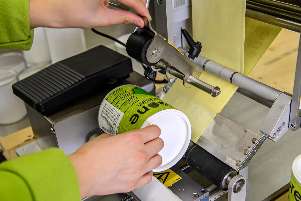 Detailaufnahme: Mitarbeiterin in einer Manufaktur verwendet eine halbautomatische Etikettiermaschine, um zylindrische Dosen zu etikettieren